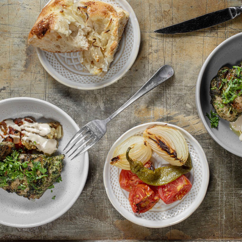 A selection of plates with focaccia from the taboon, grilled antipasti vegetables, a piece of Zaatar chicken accompanied by roasted eggplant