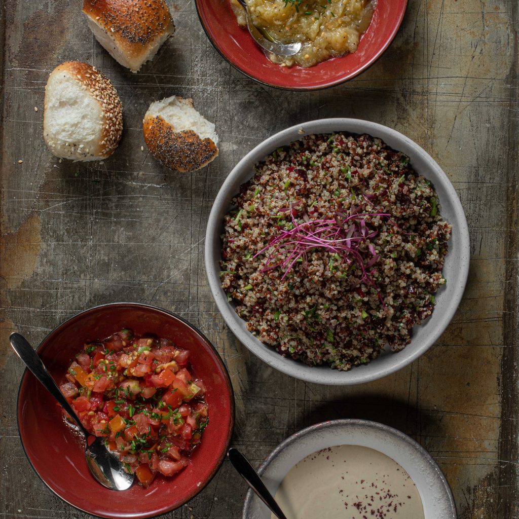 Serving bowls with quinoa and cranberry salad, cherry tomatoes salad and grilled eggplant salad accompanied by mini challah bread