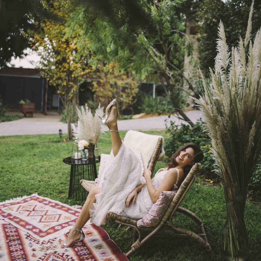 A bride sits and raises her leg on a bamboo chair in a styled erea on the grass at the reception in the weddings garden
