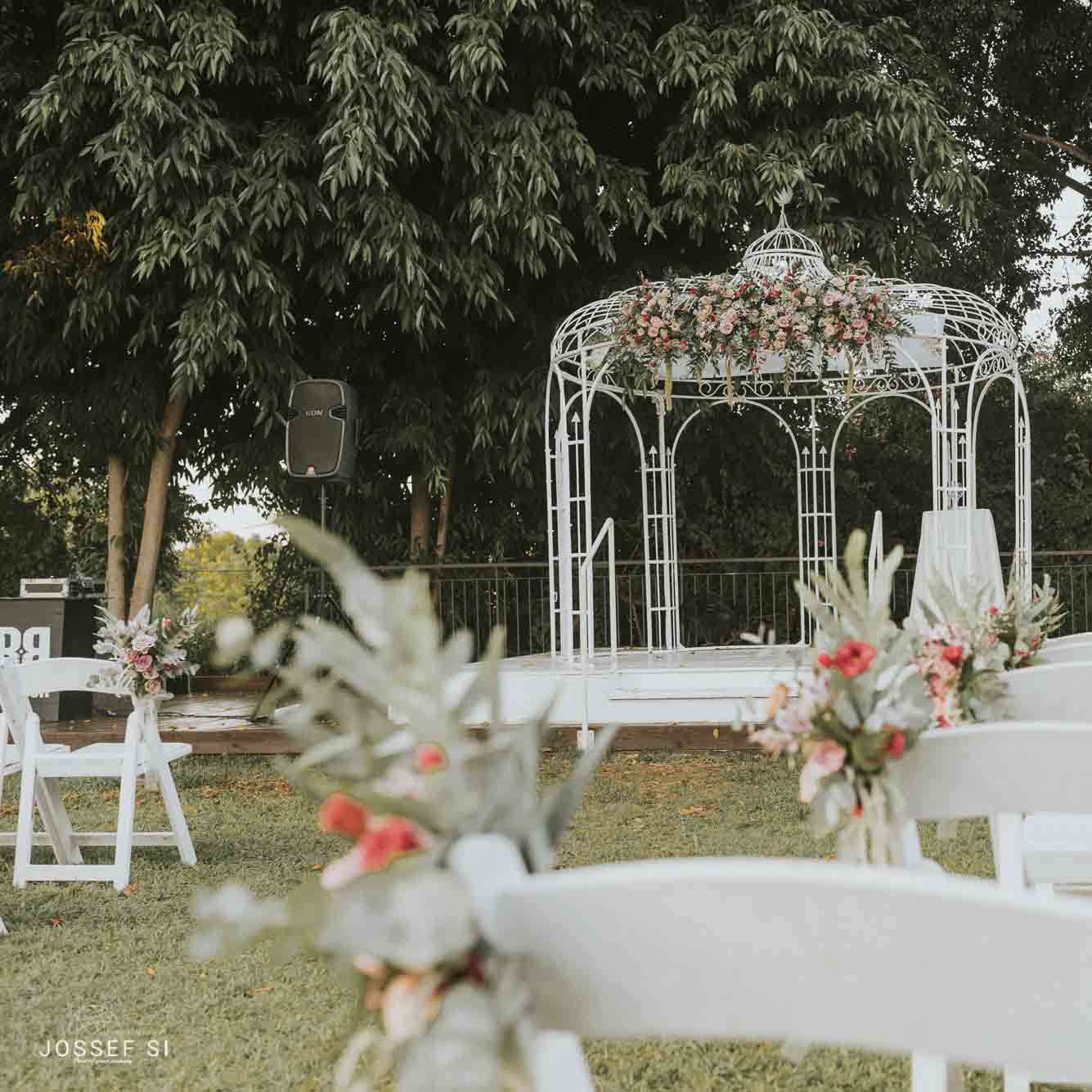 גזיבו לבן מקושט בפס פרחים וזרי פרחים קטנים קשורים לכסאות עץ לבנים על הדשא