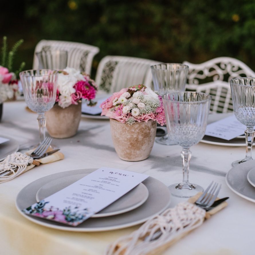 A table set with a tablecloth, plates, a menu on each plate, cutlery in a macrame holder, wine glasses and clay pots with flowers