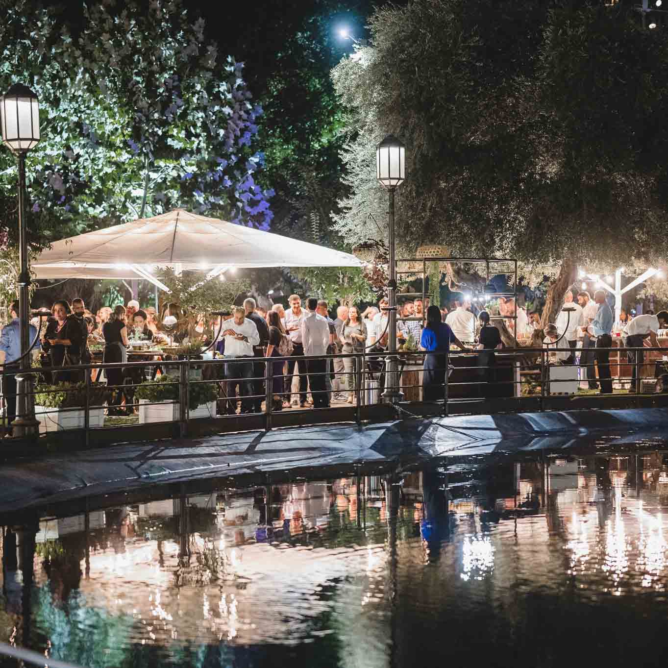 Guests at a reception in the garden under trees and an illuminated white parasols, reflected in the pool water