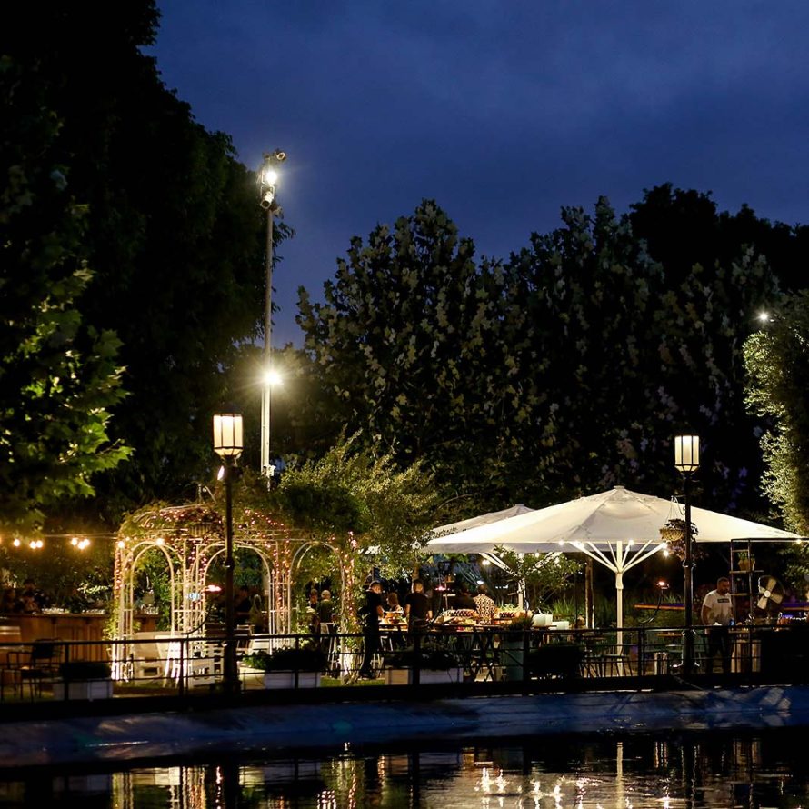 A water pool , a lighted gazebo, white parasols, lit street lamps and old trees