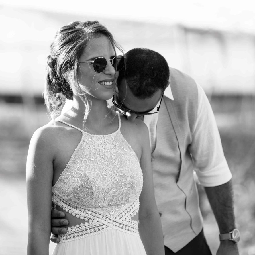 Groom hugs and kisses the brides’ shoulder who is wearing sunglasses