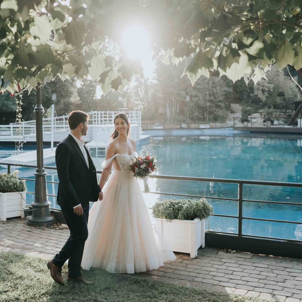 Ronell & Guy חתן וכלה צועדים בשביל מרוצף המקיף את בריכת המים בגן האירועים