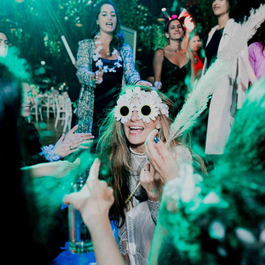 A bride with flower-shaped glasses smiles and surrounded by friends on the dance floor