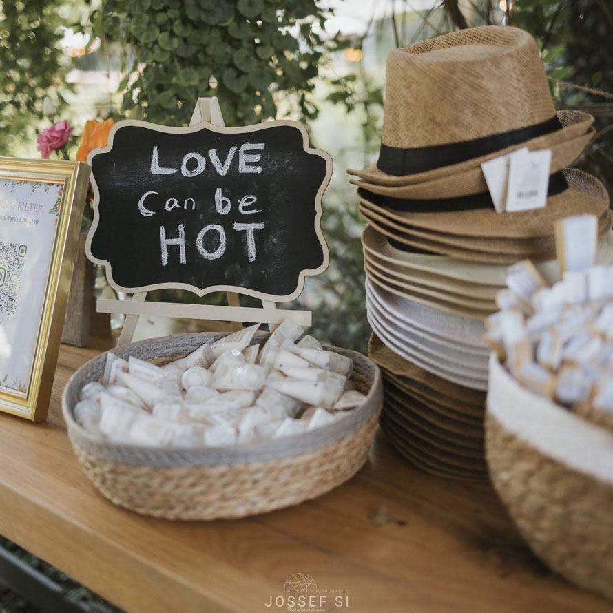 2Q4A1970--jossef-si עמדת קבלת פנים מעוצבת עם כובעי קש לאורחים, סלסילות אביזרים ושלט קטן עם הכיתוב “Love can be hot”. פינה כפרית ומוקפדת שמוסיפה טאץ’ אישי לאירוע