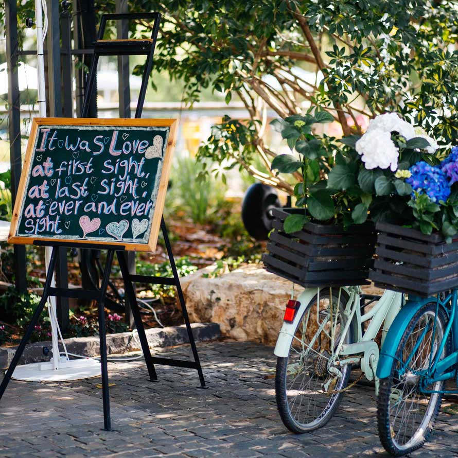 Bicycles with flower baskets next to a sign on a black stand at the entrance to the weddings garden
