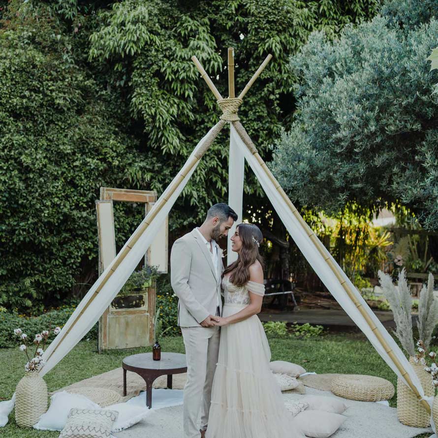 Bride and groom under a designed bamboo Teepee on the grass