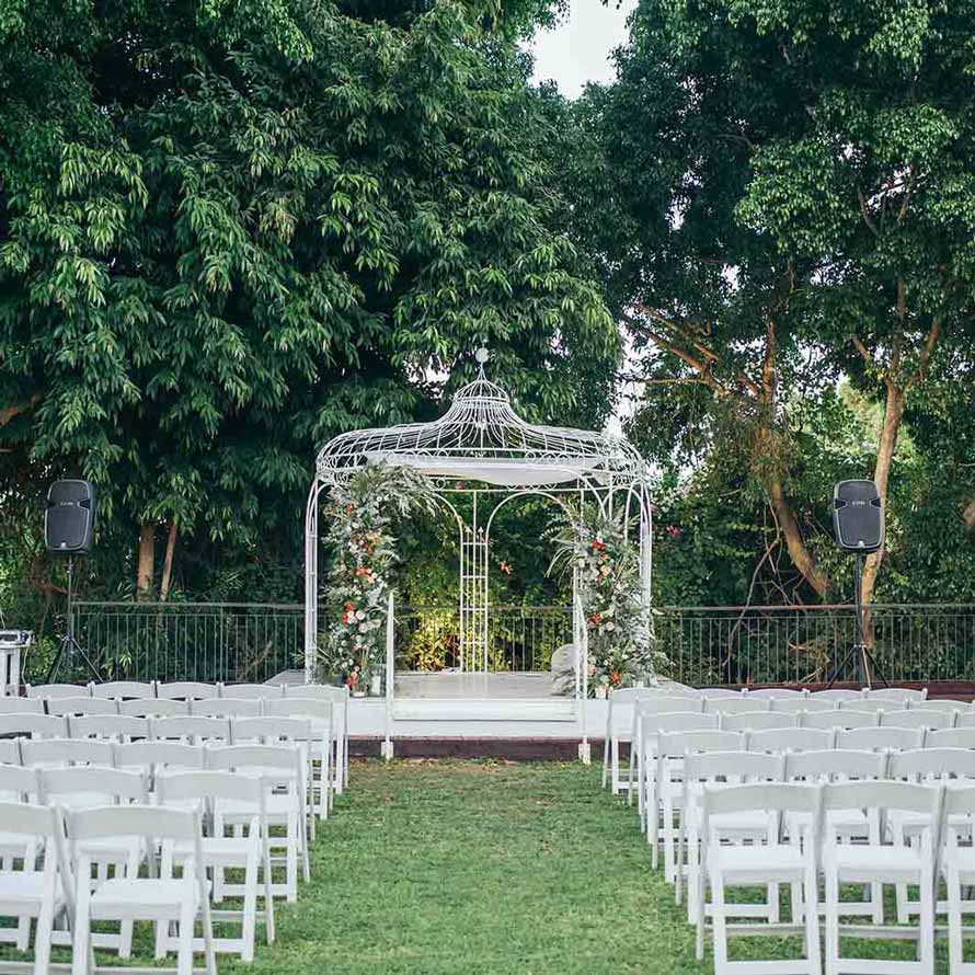 A gazebo canopy is decorated with flowers and white wooden chairs are arranged on the grass in front of the canopy