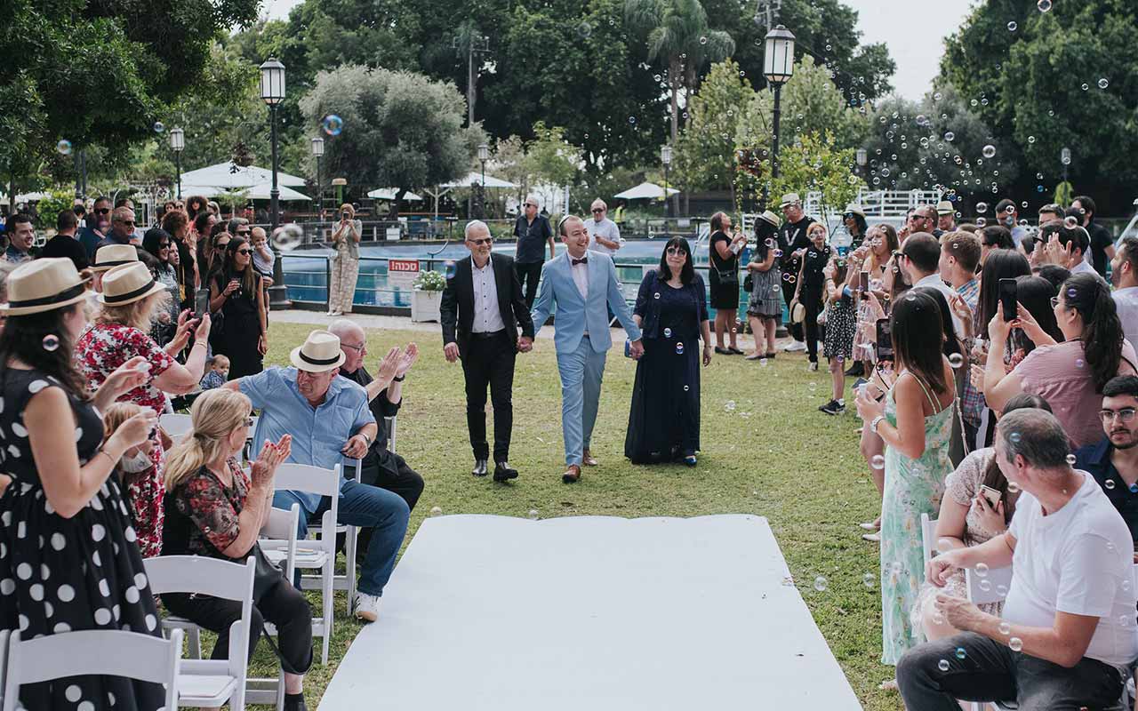 אורחים מפריחים בועות סבון ומוחאים כפיים לכבוד חתן המלווה בהוריו צועד לחופה