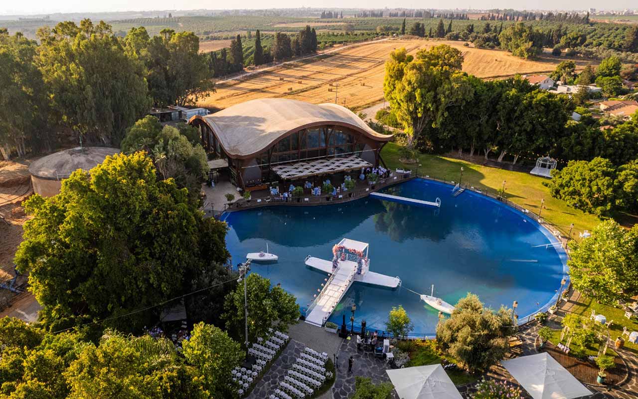 מבט מלמעלה על גן האירועים: אולם אירועים דמוי פגודה על שפת מאגר מים כחול עגול, מוקף מדשאה ועצים. ברקע שדות ופרדסים
