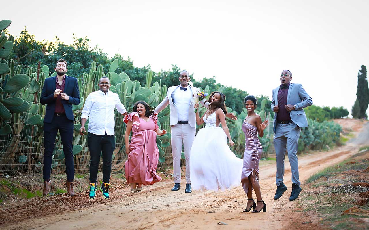 חתן וכלה ומלוויהם קופצים בשביל ליד גדר קקטוסים