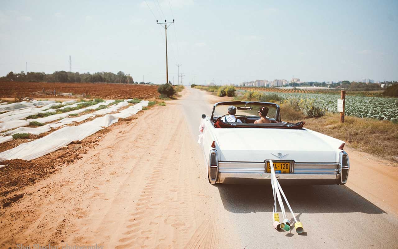 חתן וכלה נוסעים ברכב אספנות לבן עם זנב פחיות בכביש בין שדות