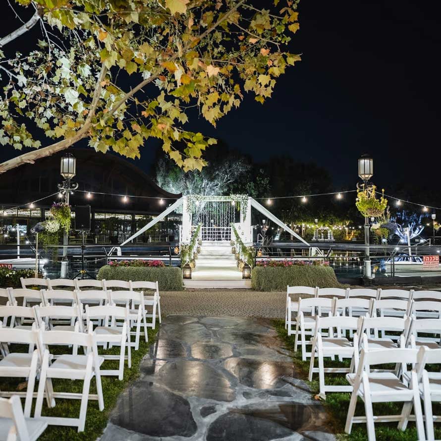 Alex&David-Weddings מתחם חופה מואר בלילה על גשר מעל המים, עם שביל אבן המוביל בין שורות כיסאות לבנים אל החופה המעוצבת. תאורת שרשראות ונרות יוצרת אווירה רומנטית על רקע האגם והאולם המואר.