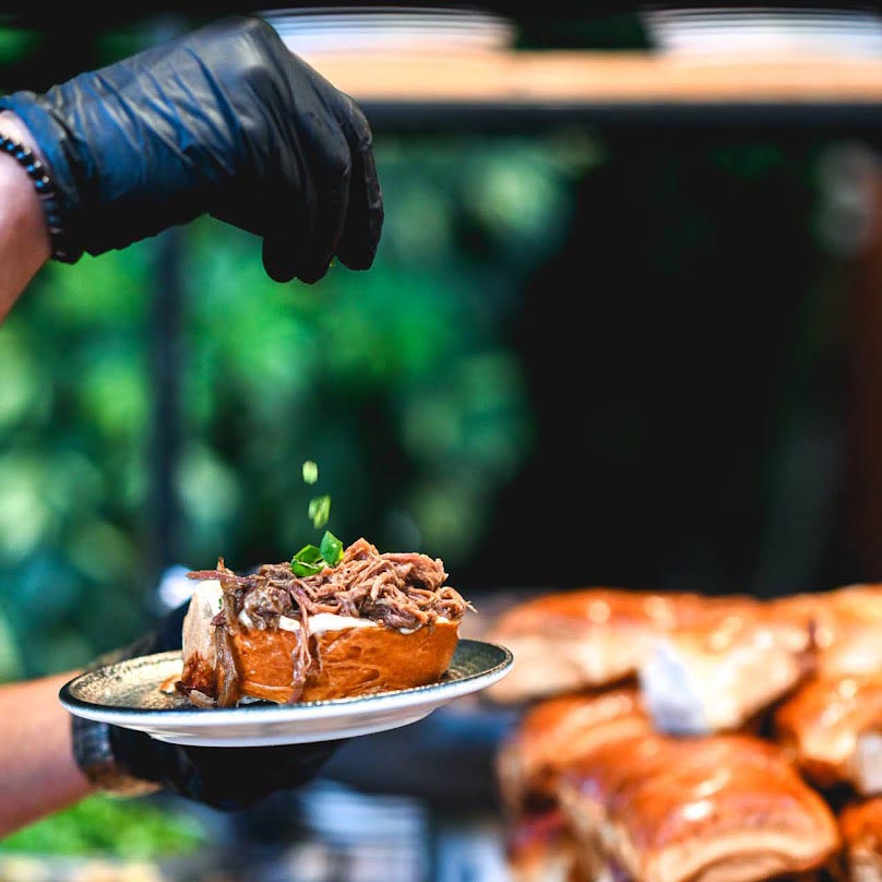 GHS_5069 מנה חמה המוגשת בצלחת אישית – לחמנייה רכה במילוי בשר מפורק עסיסי, כשיד בכפפה שחורה מפזרת עשבי תיבול טריים מעל.