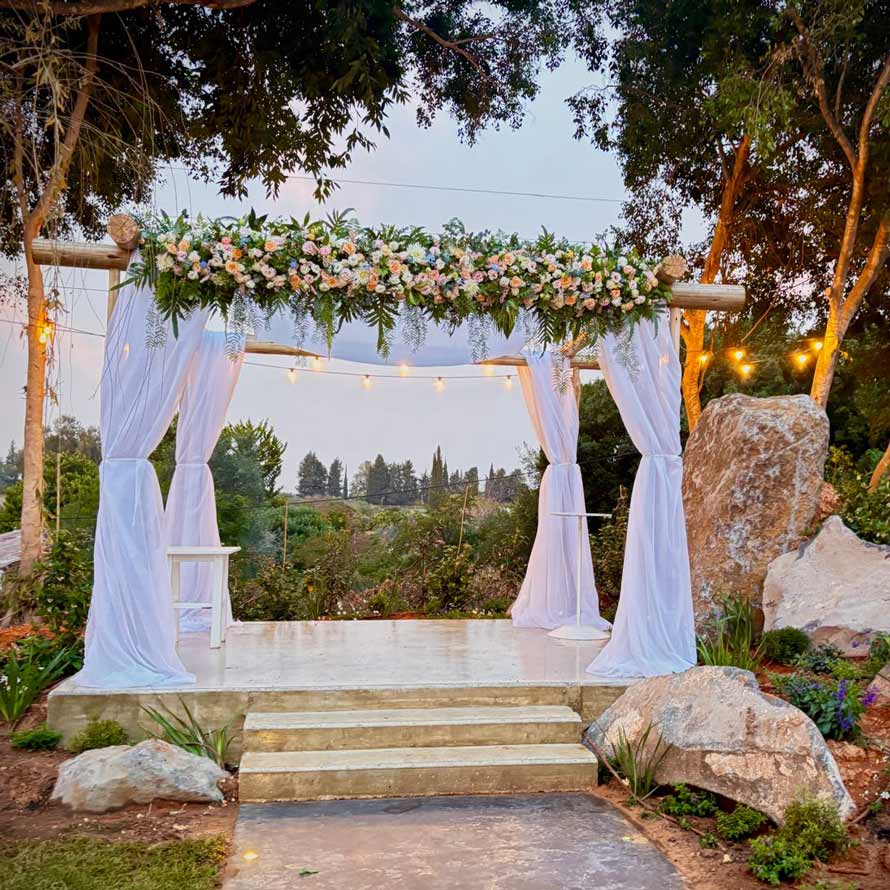 IMG-20260212-WA0002 חופה לבנה מעוצבת עם בדים לבנים רכים וסידור פרחים עשיר בגווני ורוד, שמנת וירוק. החופה מוצבת על במה קטנה מוקפת עצים וסלעים טבעיים, באווירה כפרית ופסטורלית.