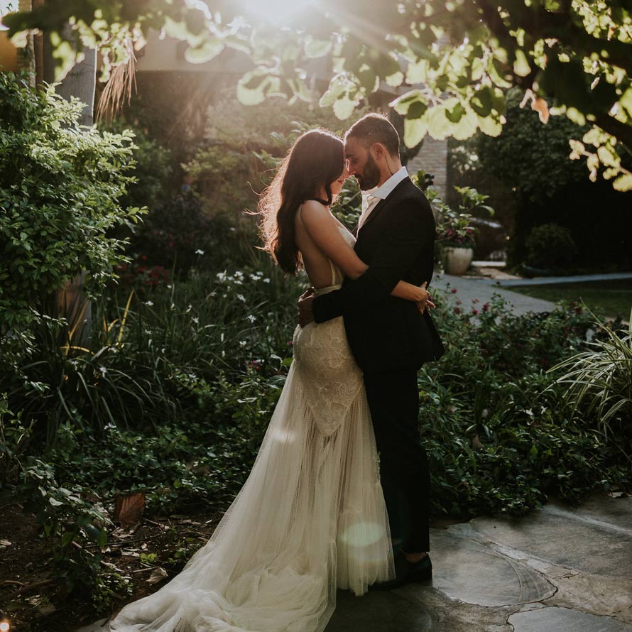 הכנותוצילומיזוג(355)שחר-וין חתן וכלה מחובקים בגן האירועים בשעת שקיעה, מוקפים צמחייה טבעית ואור רך שחודר בין העלים- רגע אינטימי ורומנטי לפני תחילת החגיגות.