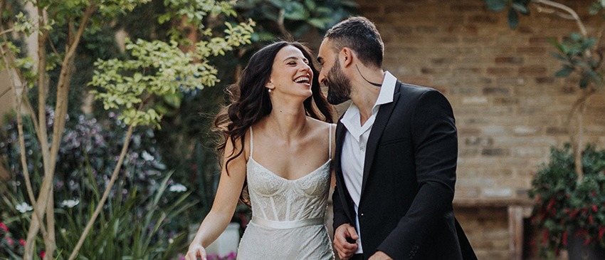 הכנותוצילומיזוג(379)שחו-וין חתן וכלה צועדים יחד בשביל אבן מוקף צמחייה ירוקה, מחייכים זה לזה באווירה קלילה ורומנטית. הכלה בשמלת תחרה בהירה והחתן בחליפה שחורה, ברגע טבעי ואינטימי בלב הגן