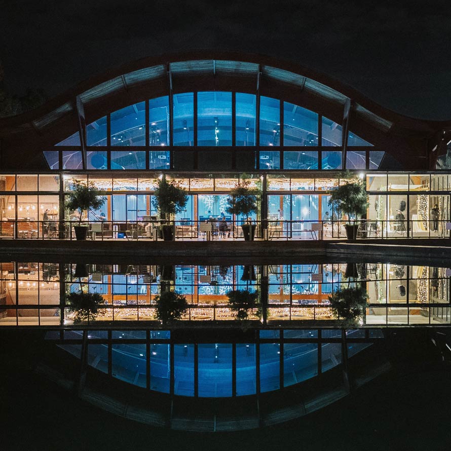 רובי-אהרון22 חזית אולם אירועים עם גג מקומר וקירות זכוכית רחבים, מוארת בלילה באור כחול וחם. המבנה משתקף במים שקטים בחזית, ליצירת מראה סימטרי ומרשים