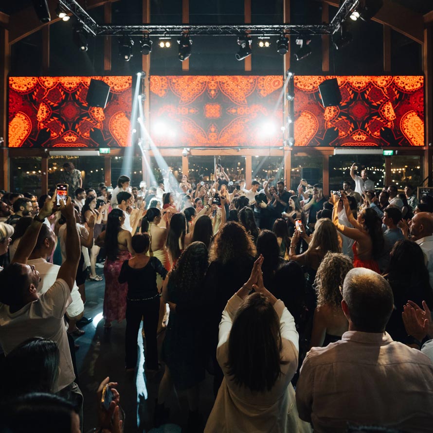 ריקודים(247)שחר-וין רחבת ריקודים מלאה באורחים חוגגים ומרימים ידיים, מוקפים תאורה צבעונית ומסכי לד דקורטיביים. אווירה אנרגטית ושמחה בשיאה בלב האולם.