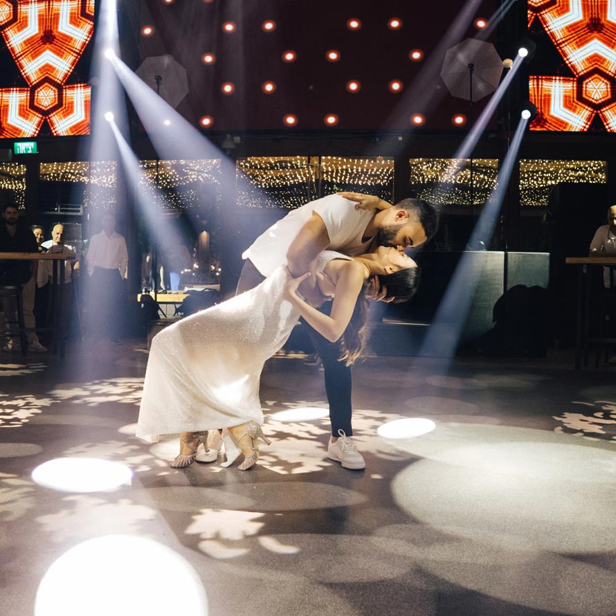 ריקודים(364)שחר-וין חתן וכלה בריקוד ראשון דרמטי במרכז הרחבה, כשהחתן מטה את הכלה לאחור בנשיקה תחת אלומות אור ממוקדות ותאורת לד חמה ברקע. רגע עוצמתי ומלא תשוקה מול האורחים.