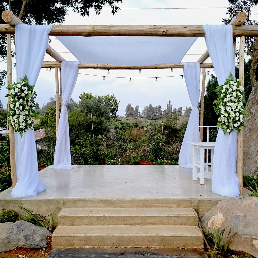 תקריב של חופה מעץ עם בד לבן וזרי פרחים, הממוקמת על במה פשוטה ומשקיפה לנוף ירוק וטבעי.