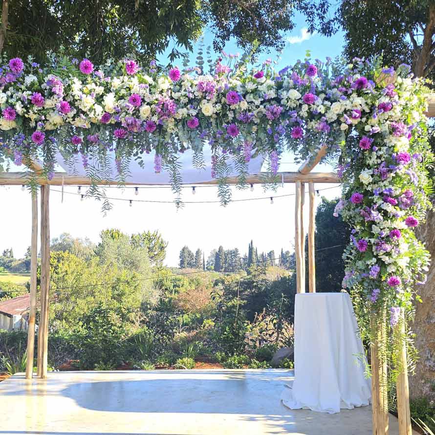 חופה מעץ מקושטת בשפע פרחים בגווני סגול, לבן וירוק, עם צמחייה משתפלת מלמעלה. החופה ניצבת על במה מול נוף טבעי פתוח של עצים ושדות, באווירה רומנטית ופרחונית באור יום.