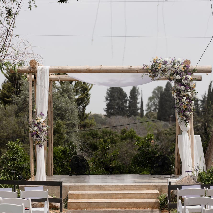AdrianbyISOGroup(255of1138) חופה כפרית מעץ עם וילונות לבנים וסידורי פרחים בגווני סגול, לבן וירוק. החופה ניצבת מול נוף טבעי של עצים וצמחייה, עם שביל גישה ומקומות ישיבה לטקס חתונה באוויר הפתוח.